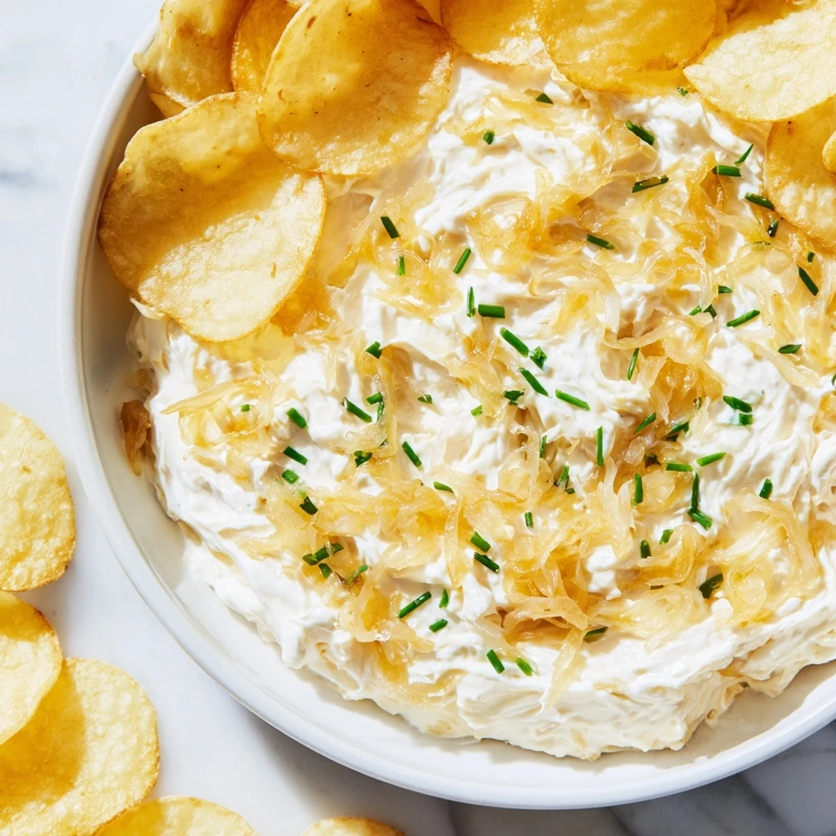 A savory homemade onion dip garnished with fresh chives, alongside a pile of thick-cut potato chips on a marble surface.