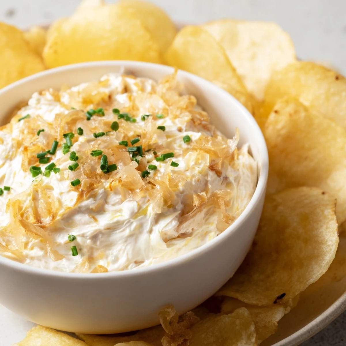 Creamy onion dip with caramelized onions in a white bowl, served with crispy golden potato chips for dipping.