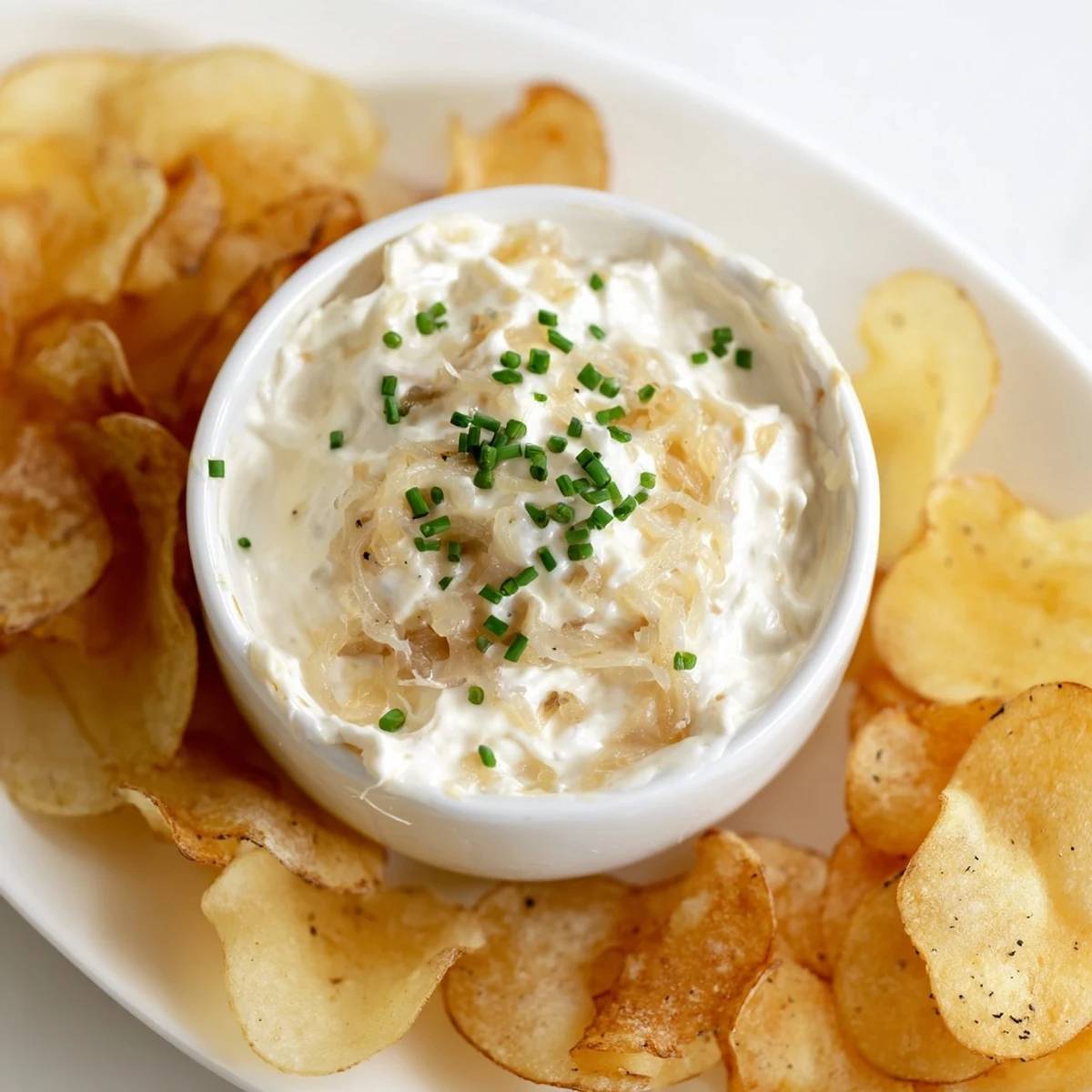 Homemade onion dip garnished with fresh chives paired with crispy russet potato chips on a white plate.