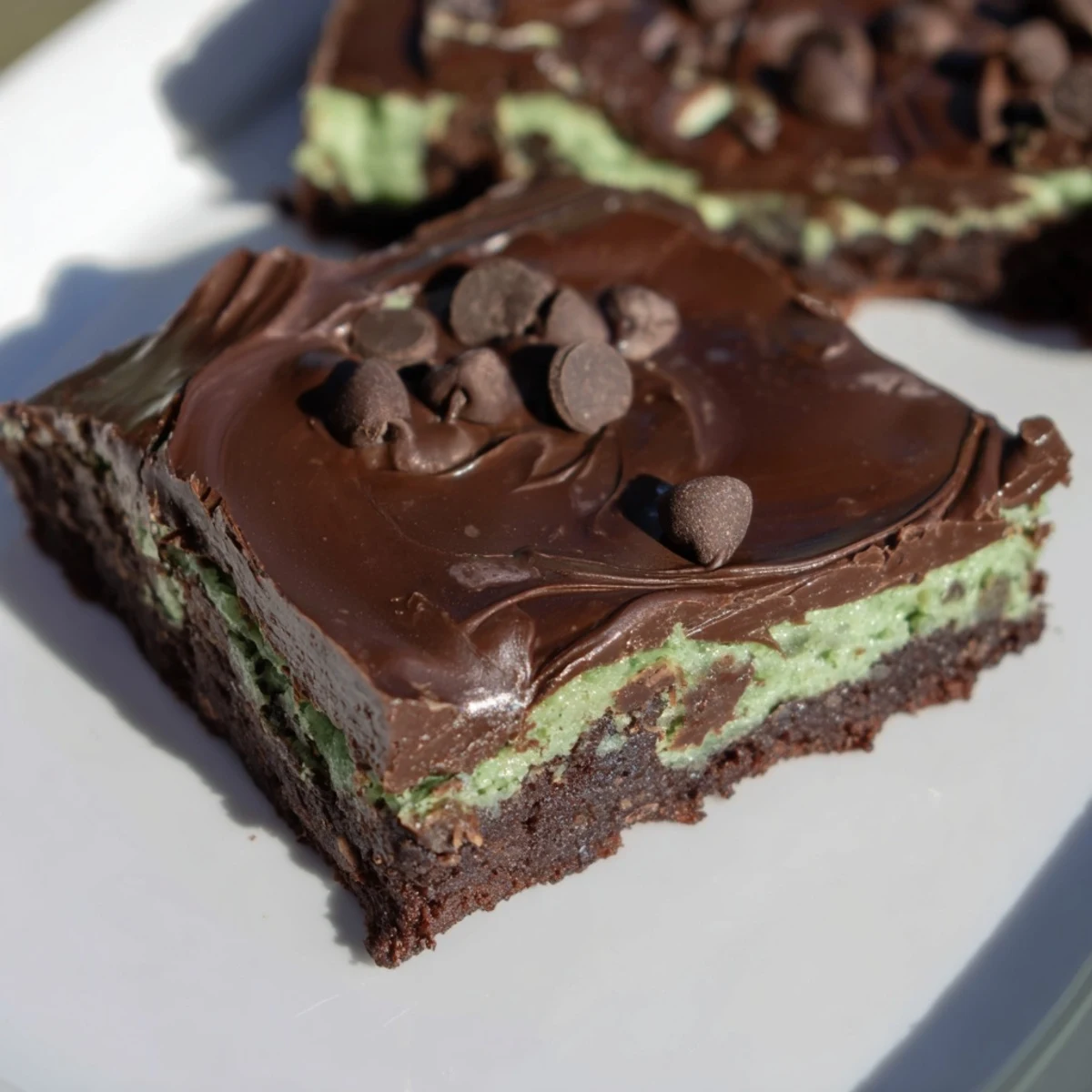 A square of Green Mint Brownies with Chocolate Chips on a white plate, showcasing a rich chocolate ganache topping and a vibrant mint swirl inside.