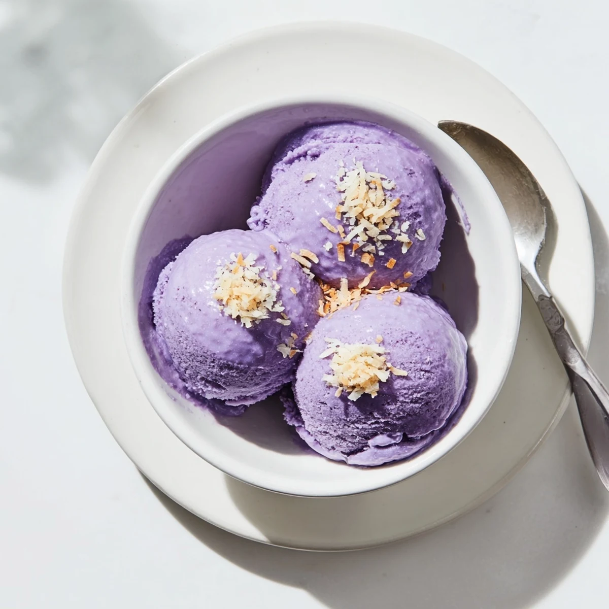 A close-up of vibrant purple ube ice cream scoops in a chilled glass bowl with toasted coconut flakes on top.