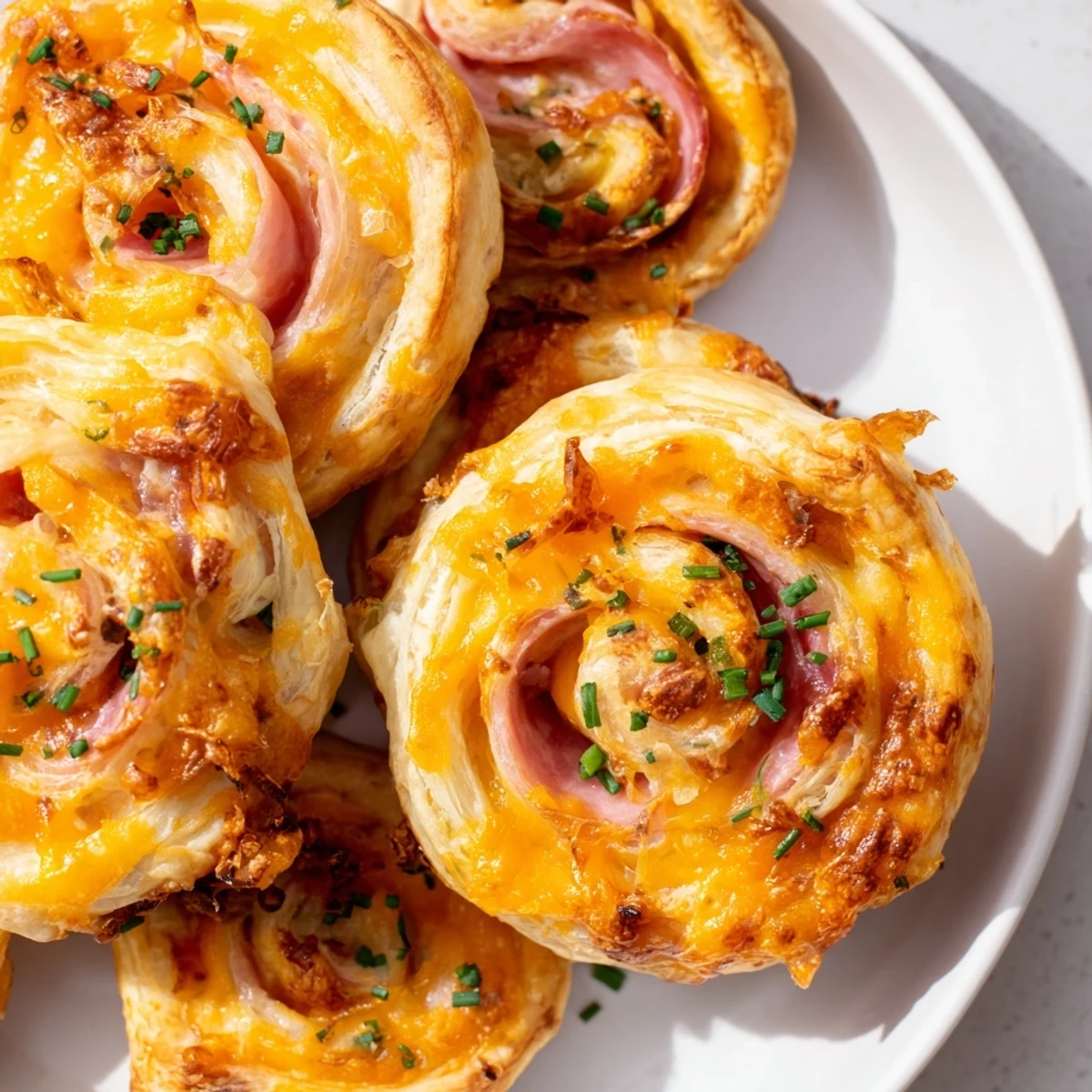Golden-baked Easter Ham Cheddar Pinwheels rest on a white plate beside fresh chives and a small bowl of Dijon mustard.