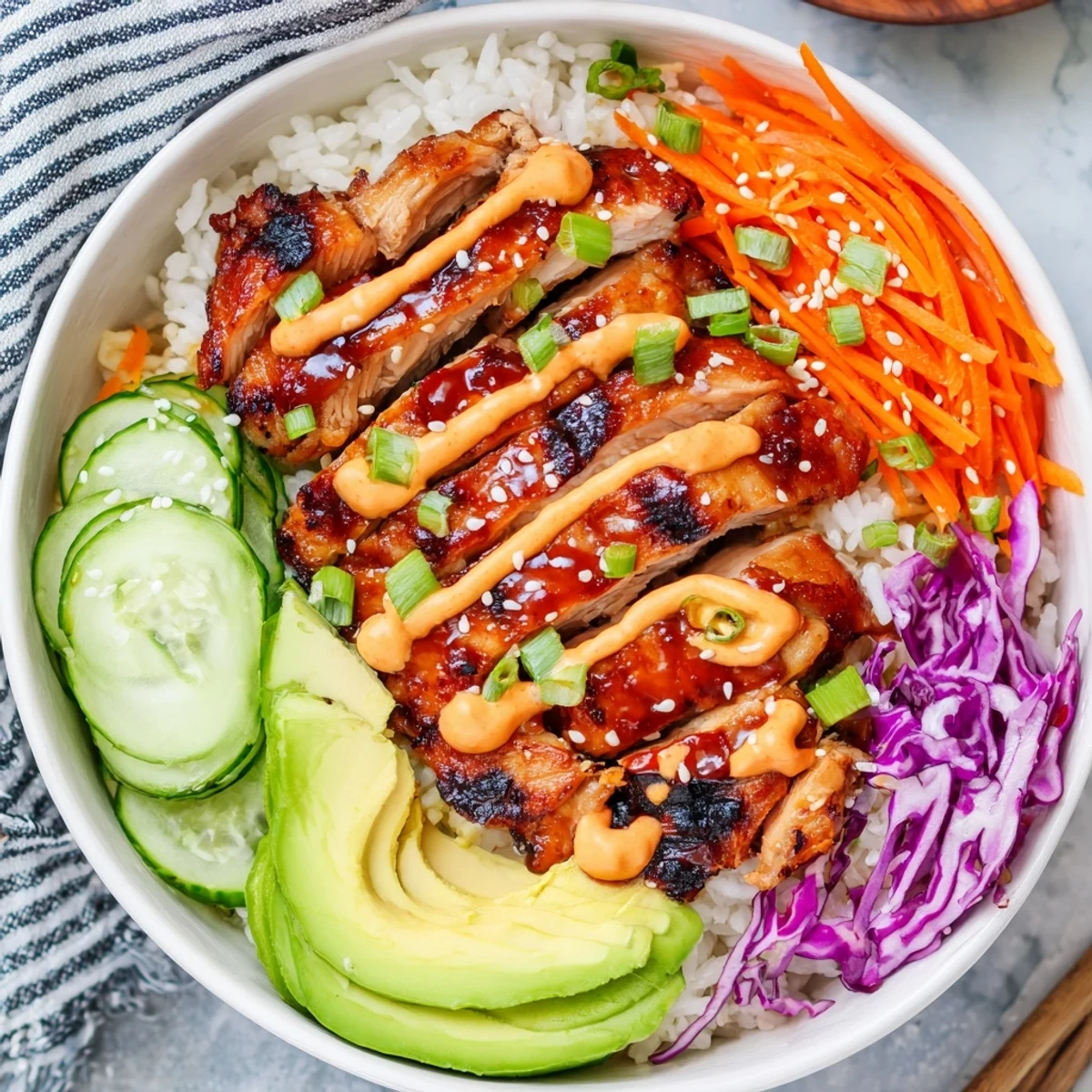 Sliced Korean BBQ chicken thighs arranged over a bed of rice with shredded carrots, cucumber, purple cabbage, and ripe avocado