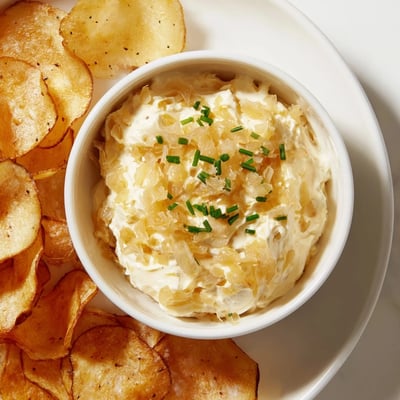 A rustic wooden board displays a bowl of savory onion dip and a pile of warm, seasoned potato chips.